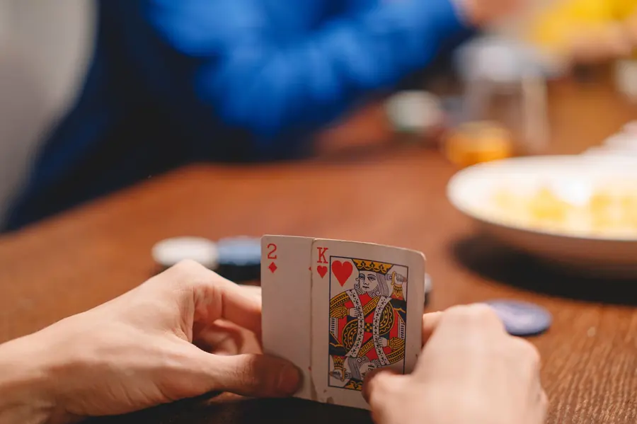 A pair of hands holding two playing cards, a two of diamonds and a king of hearts, over a wooden table with chips and a plate out of focus behind
