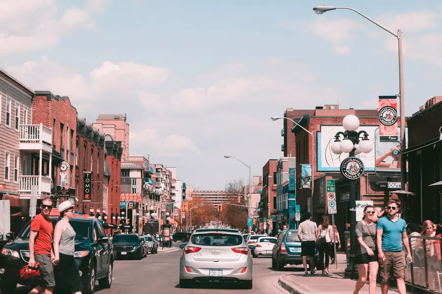 A busy downtown street in a Canadian city, with brick low-rise buildings, shops, parked cars and pedestrians on a sunny afternoon