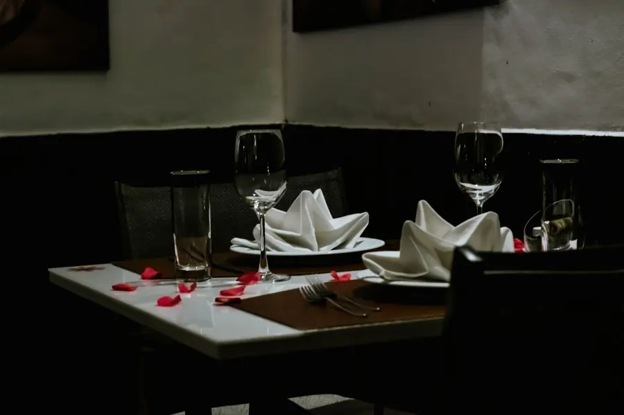 A dimly lit restaurant table set for two, with folded white napkins, wine glasses and scattered red petals on a dark tablecloth