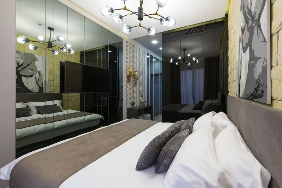 A modern hotel bedroom with a large bed, white and grey linen, a mirrored wall, a wall-mounted television and a globe-light chandelier