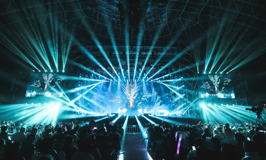 A packed arena concert seen from the back of the floor, with teal and white beams from overhead stage lighting fanning out above the crowd